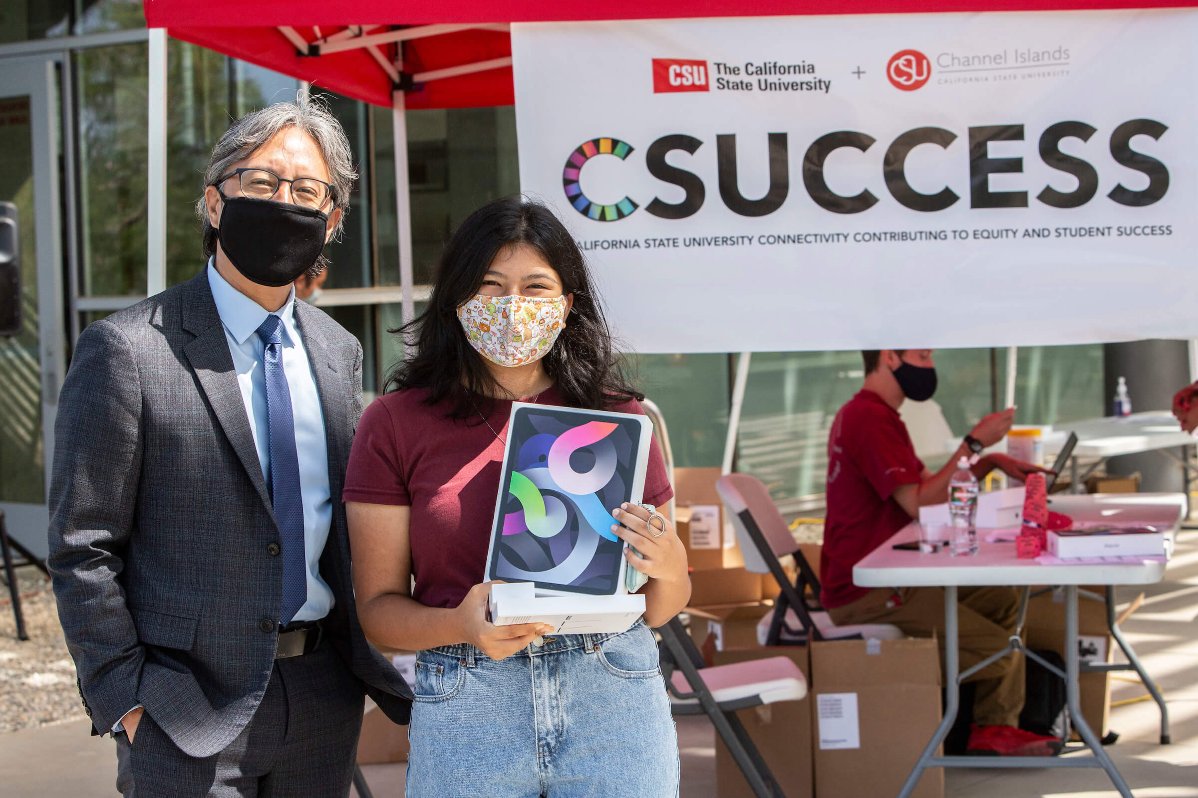 man stands next to woman holding a new iPad in a box outside on a college campus. 