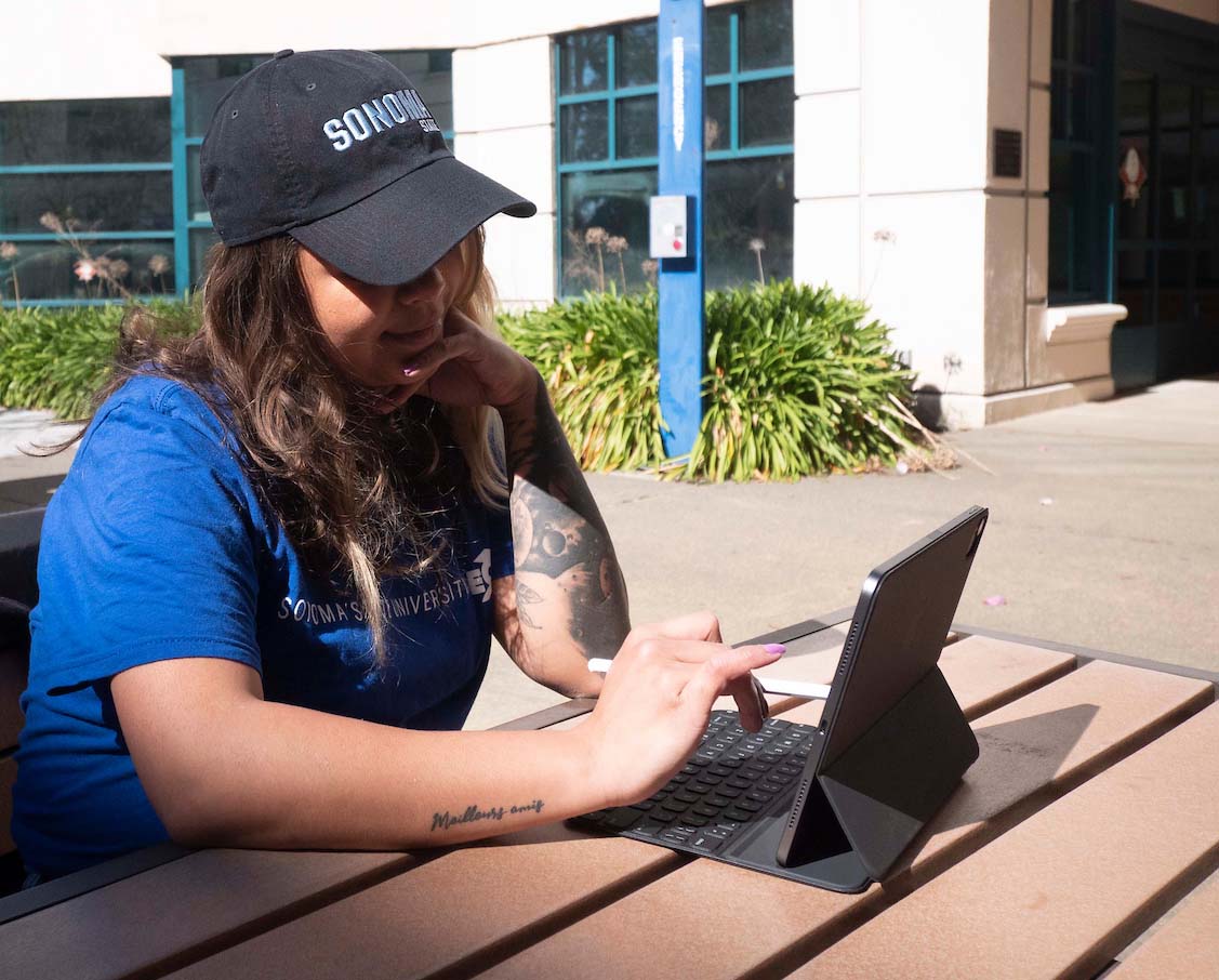 student with long hair and cap sits at outdoor table typing on a tablet with keyboard