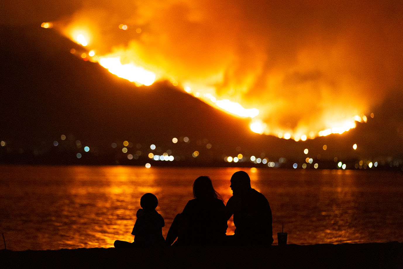 people sitting by a lake looking at mountains on fire at night