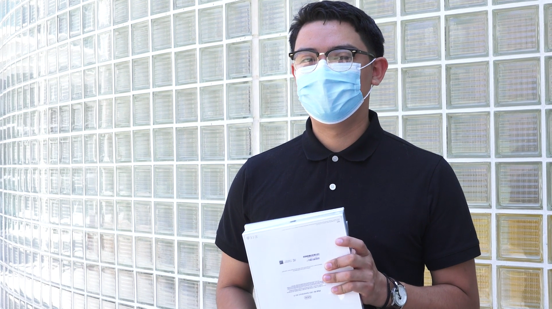 young man in polo shirt and protective face mask stands holding white boxes.