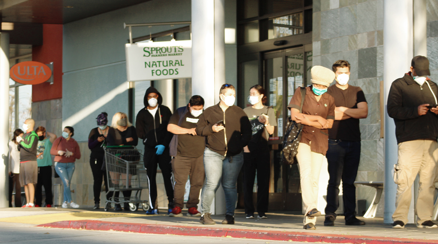 People wearing masks stand in line to enter Sprouts Farmers market grocery store.