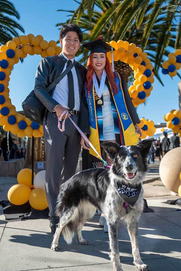 graduating student and her guest pose with a dog whose bandana says my mom did it
