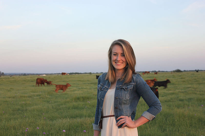 Grace Woodmansee wearing a white dress and denim jacket smiling in front of a field with several brown cows