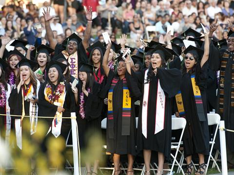 A large group of people wearing graduation caps and gowns cheering and smiling.