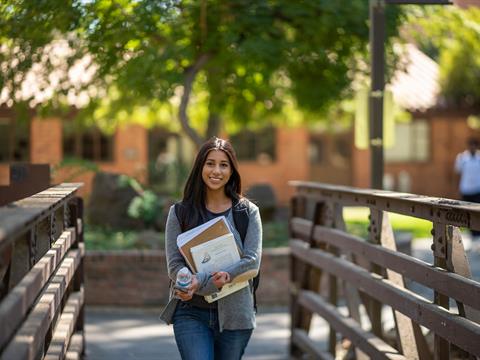 female college student walks across bridge holding books on campus